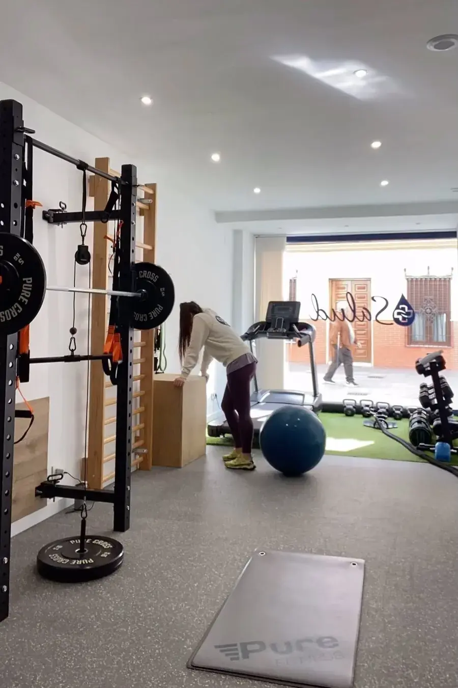 Sala de entrenamiento de +Salud con rack de fuerza, cinta de correr y el logo en el escaparate